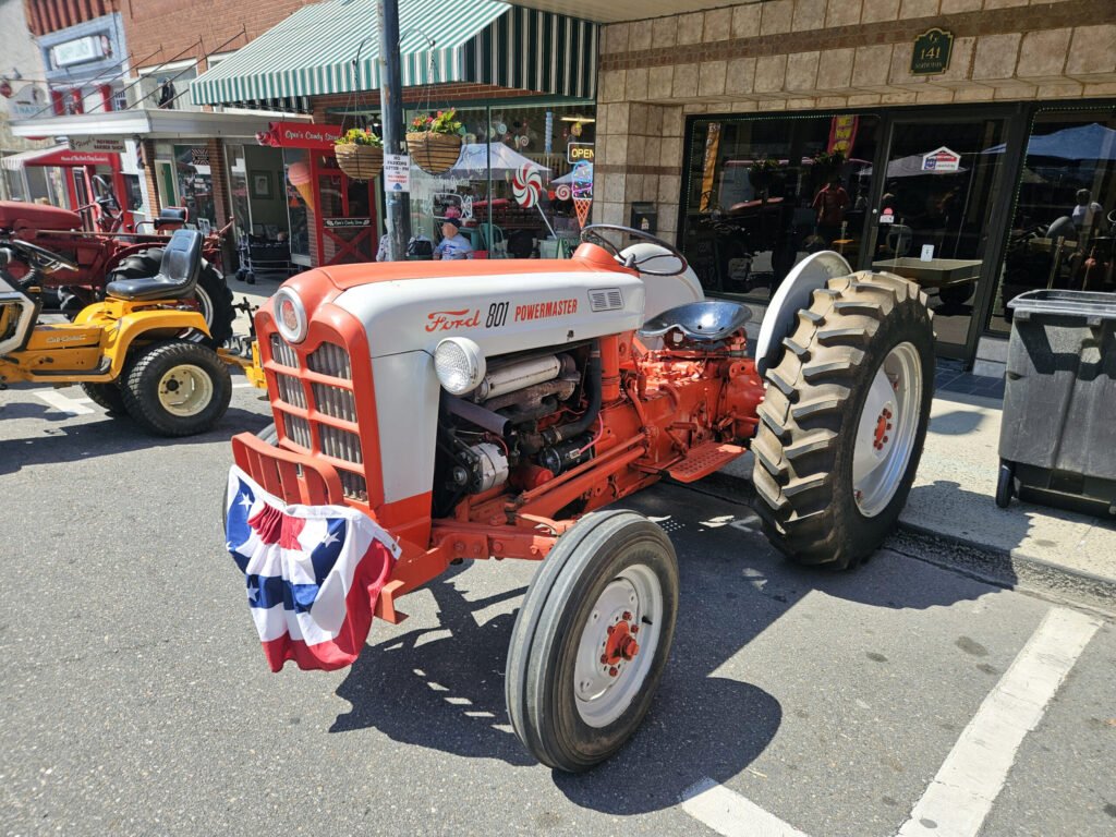 Red and white Ford 801 Powermaster tractor parked on a city street with storefronts and striped awnings in the background