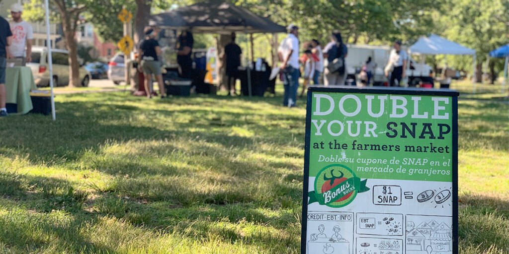 Outdoor farmers market scene with a large green promotional sign in the foreground that reads 'DOUBLE YOUR SNAP at the farmers market,' shoppers and stalls visible under tents in a sunny park setting.