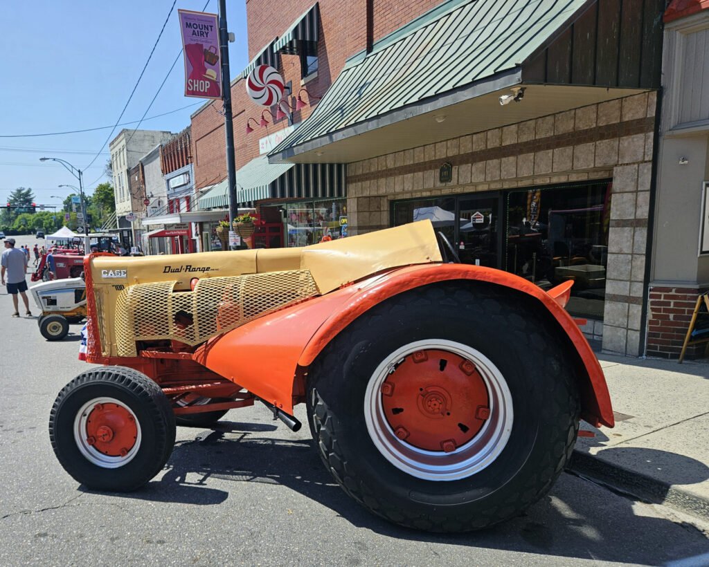 Orange tractor with large rear wheel parked on a sunny downtown street with brick storefronts in the background.