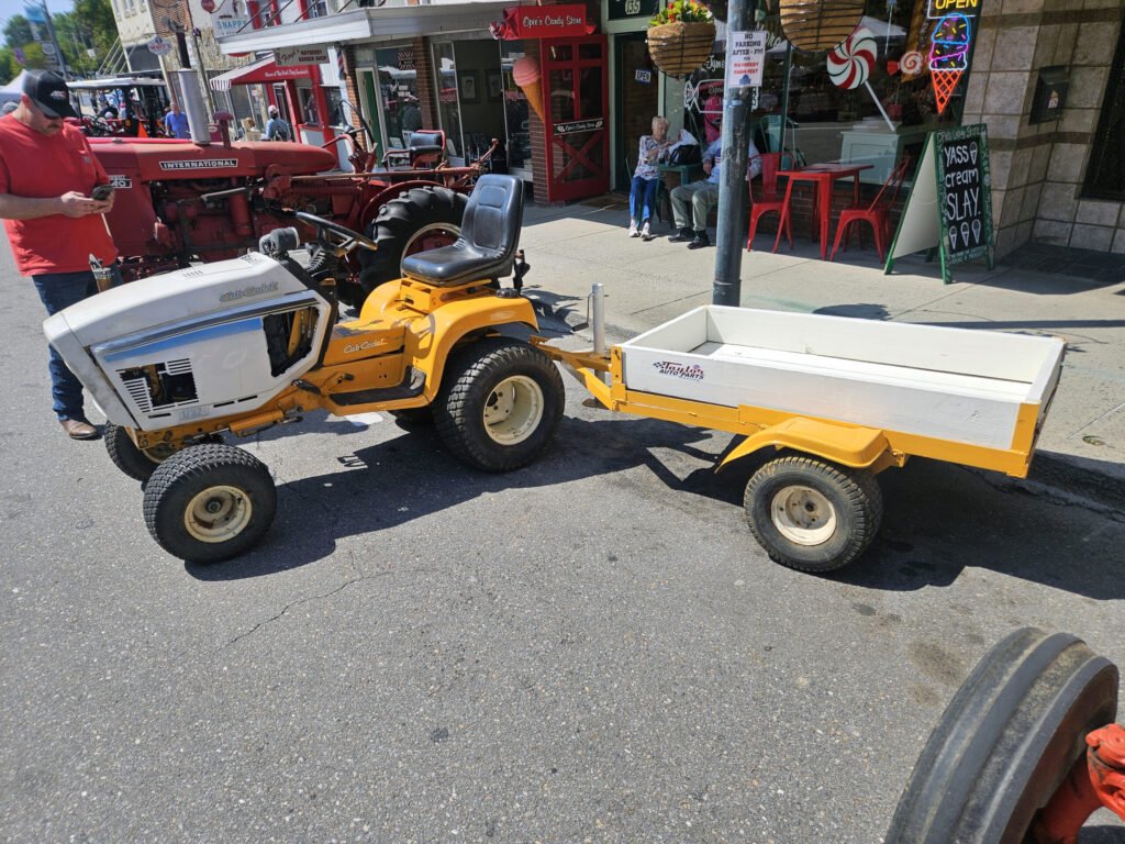 Man in a red shirt stands beside a red International tractor pulling a yellow trailer on a busy downtown street with shops and pedestrians in the background.