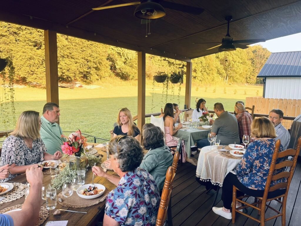 Group of people dining at a long wooden table under a covered patio, autumn sunlight in the yard beyond the railing.