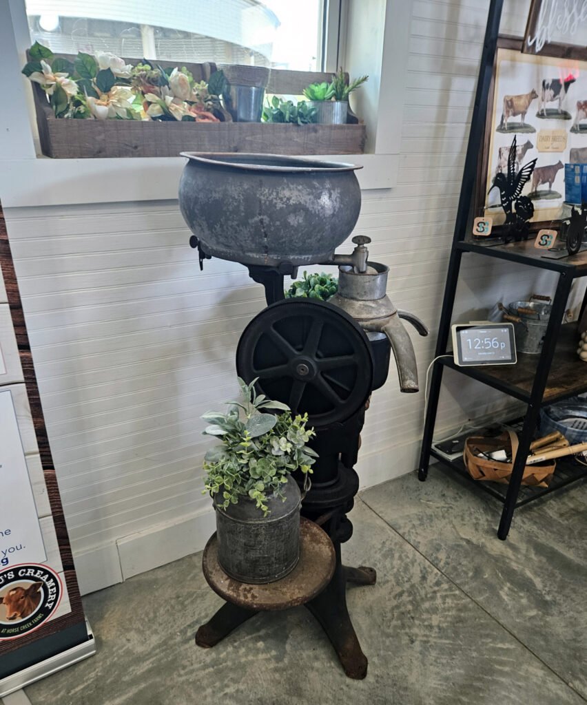 Antique metal grinding mill on a stand with a wheeled wheel, topped by potted greenery, in a store display.