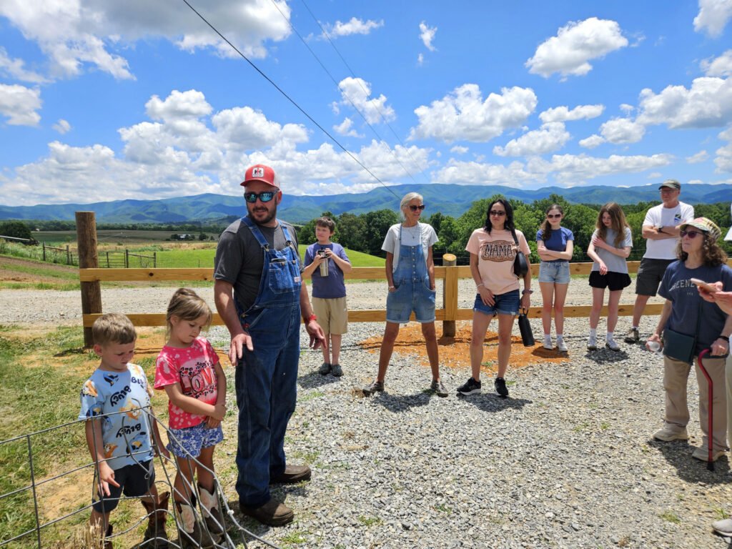 Group of adults and children standing along a wooden fence outdoors on a sunny day, with mountains and blue sky in the background.