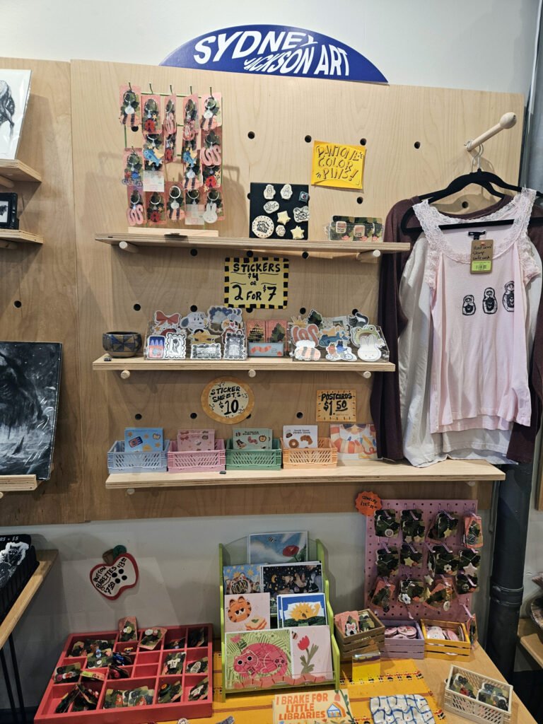 Wooden pegboard display with colorful stickers, pins, and small cards arranged on shelves, plus a pink shirt on the right.