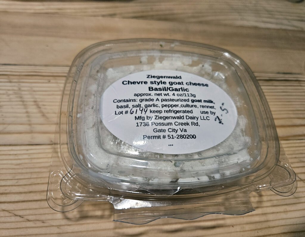 Plastic container of Chevre-style goat cheese labeled Basil/Garlic on a wooden table; shows approximate 4 oz (113 g) net weight and ingredients list.
