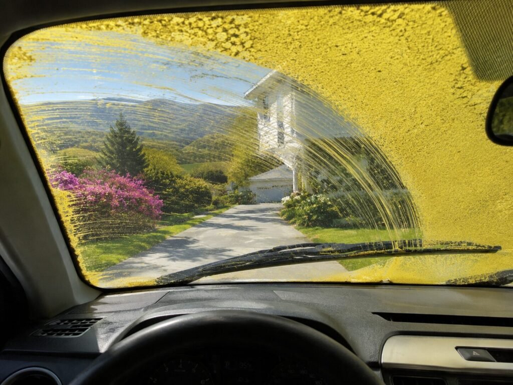 Inside a car during a wash, the windshield is covered in yellow soap streaks while a sunny driveway, flowering bushes, and a white house appear beyond the glass in the background at the hills.