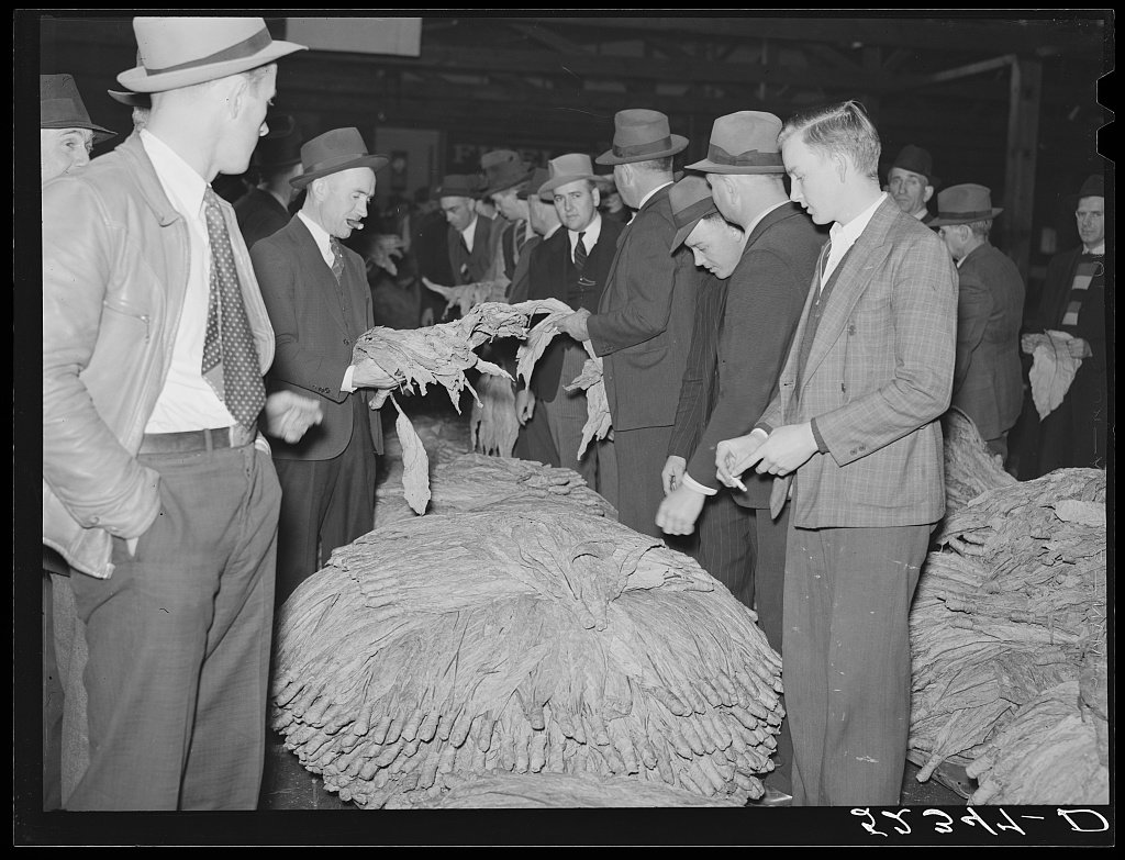 Group of men in 1940s suits and hats inspect a large, textured, feather-like display laid out on a table at an exhibition.