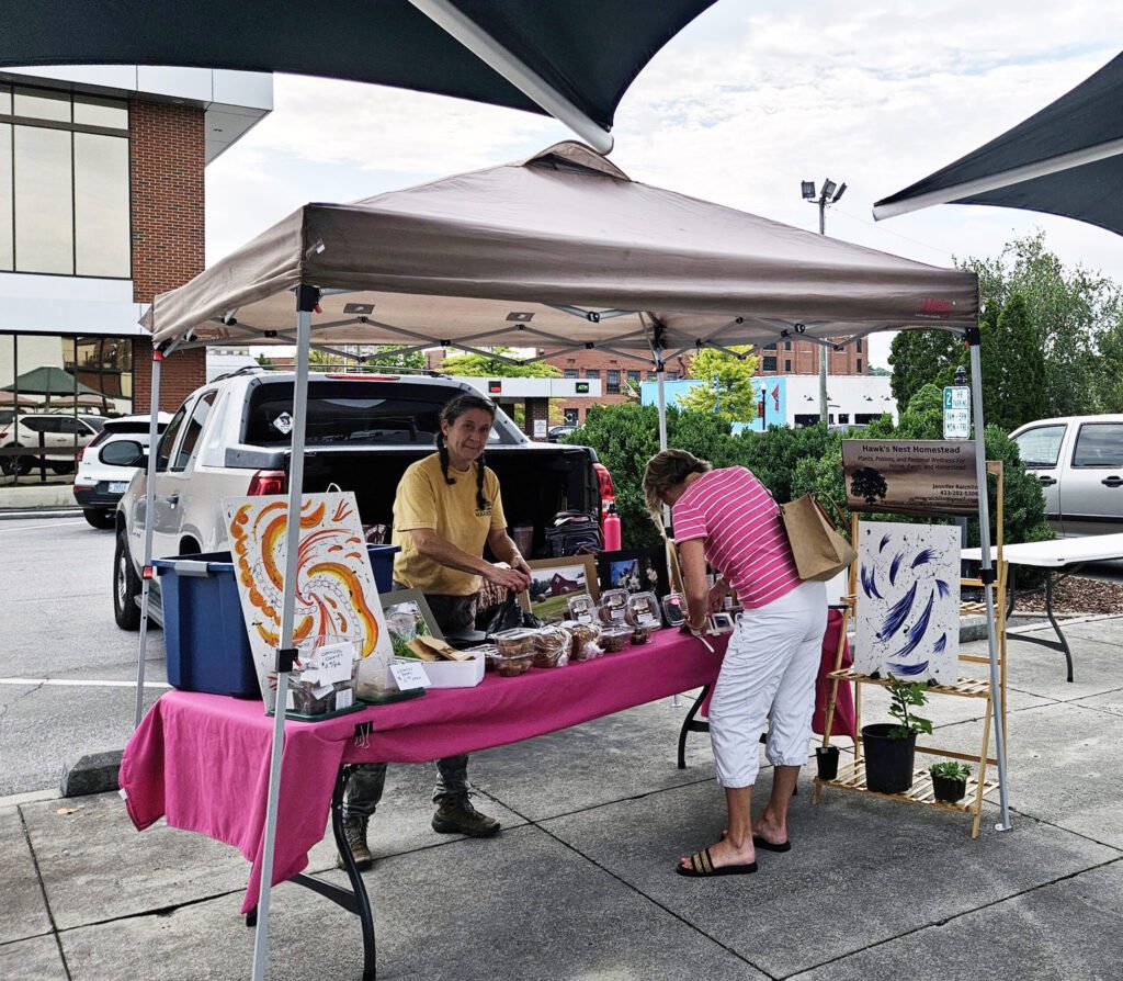 20260808_095021 - Appalachian Highlands Farmers Magazine Outdoor market stall with a pink tablecloth displaying paintings and crafts; two people are at the booth in a parking lot.