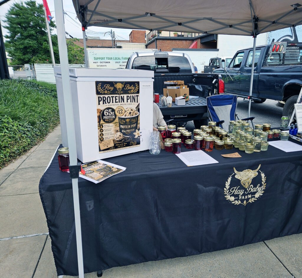 20260808_095123 - Appalachian Highlands Farmers Magazine Outdoor market stall under a canopy, displaying a'Hay Baby Farm Protein Pint' poster and rows of mason jars on the table with a pickup truck nearby.
