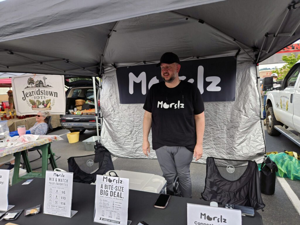20260808_111809 - Appalachian Highlands Farmers Magazine Vendor at an outdoor market stall under a grey canopy, wearing a black shirt and cap, with menu cards on the table in front of him.