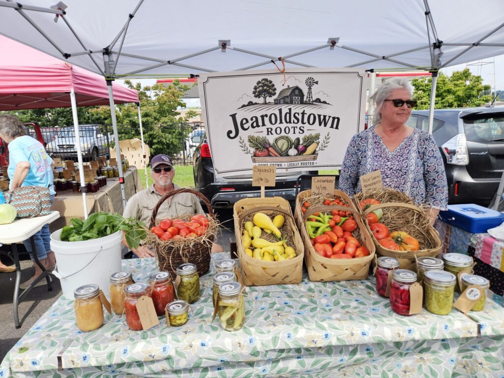 20260808_112230 - Appalachian Highlands Farmers Magazine Outdoor farmers market stall with baskets of tomatoes, peppers, and jars of preserved goods under a canopy; sign reads Jearoldstown Roots west of the setup.