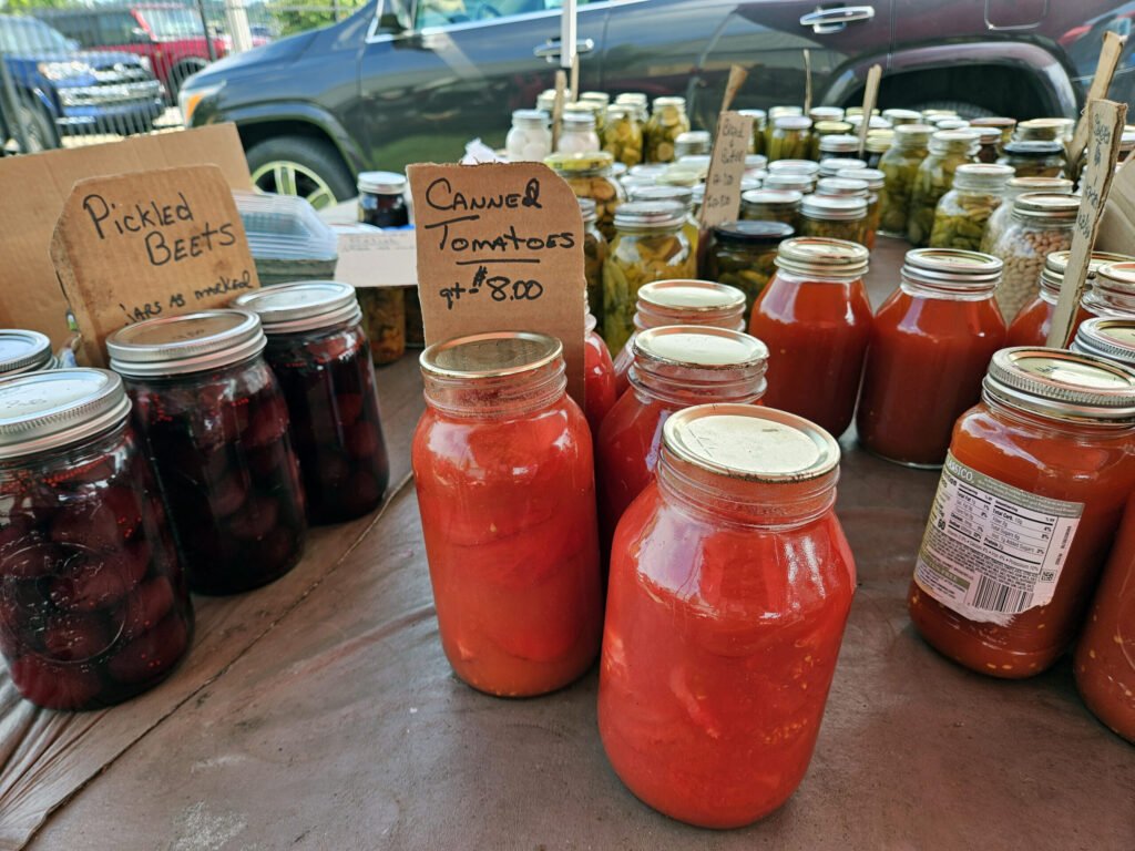 20260808_112527 - Appalachian Highlands Farmers Magazine Table filled with jars of red tomato juice and pickled produce; cardboard signs read'Pickled Beets' and 'Canned Tomatoes'