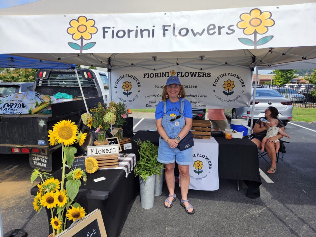 20260808_112919 - Appalachian Highlands Farmers Magazine Smiling woman vendor at a sunny flower stall under a white tent, with bright sunflowers and a Fiorini Flowers banner in the background.