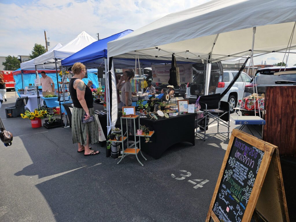 20260808_113603 - Appalachian Highlands Farmers Magazine Outdoor street market with white tents and vendors selling items; chalkboard sign in the foreground and cars in the background