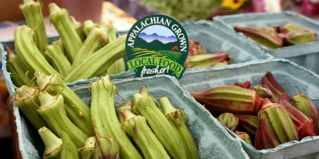 Okra pods in blue crates at a farmers market with an Appalachian Grown Local Food sticker saying'fresher!'