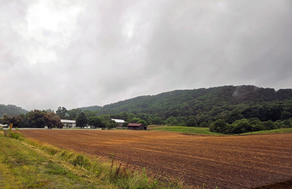 Cloudy sky over a rural landscape with a plowed field, trees, and forested hills in the distance. - Photo by A. Creighton