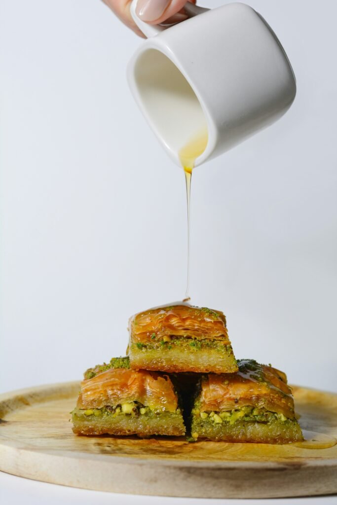 aidin-geranrekab-tQehavfgNMM-unsplash - Appalachian Highlands Farmers Magazine Hand pouring a thin stream of golden syrup onto a stack of pistachio baklava on a wooden board.